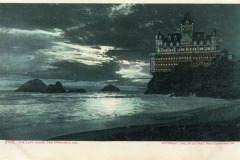 A large, illuminated Victorian-style building—reminiscent of San Francisco’s historic Cliff House—sits on a rocky cliff above the ocean at night, with waves breaking below and a dramatic moonlit sky overhead.