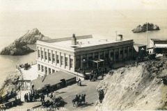Historic photo of the Cliff House in San Francisco overlooking the ocean, with people, horses, and carriages around the iconic building. Rocky outcrops and additional small structures can be seen nearby.