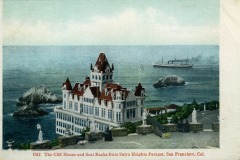 Historic postcard image of San Francisco’s Cliff House, a large, ornate building with red roofs perched on a rocky coast. Statues and parapet stand in the foreground, with a ship and rocks visible in the background overlooking the ocean.