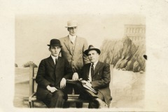 Three men in formal suits and hats pose for a vintage black-and-white photograph; two are seated on a bench and one stands behind them, with a painted seaside backdrop of San Francisco’s iconic Cliff House and rugged cliffs.