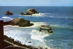 A building, reminiscent of San Francisco’s iconic Cliff House, sits partially submerged offshore amid ocean waves, with rocky outcrops in the background under a clear blue sky.