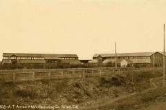 Sepia-toned photo of A.T. Ames Manufacturing Co. in Niles, California. Two large industrial buildings with long, narrow windows stand behind a wooden fence and barren land. The sky is clear, and a utility pole is visible.