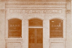 Front view of an old brick building labeled Bank of Centerville. The facade features three arched windows with the banks name printed on them. The entrance door is centered below a brick arch. The building sits on a blank street.