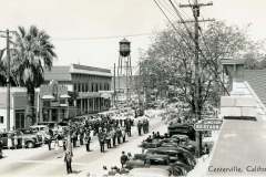 A black and white photo of a bustling street scene in Centerville, California, featuring a parade with a marching band. Vintage cars line the street, and a tall water tower is visible in the background. A Lincoln Restaurant sign is seen on the right.