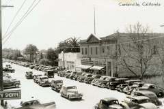 A vintage black-and-white photo of Centerville, California, showing a busy street lined with classic cars. On the right, historic brick buildings and palm trees are visible. Signs for a grocery store and a beauty shoppe are present.