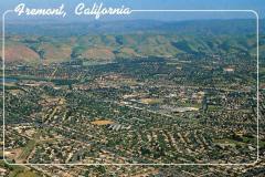 Aerial view of Fremont, California, featuring sprawling residential neighborhoods and road networks. The background showcases rolling hills under a clear blue sky, with white text labeling the location at the top left.