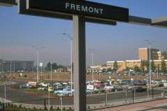 View from a BART train station platform showing a sign reading FREMONT taken in October 1974. Below is a parking lot filled with vintage cars. In the background, a multi-story building is visible under a clear blue sky.
