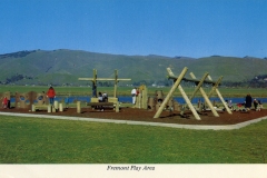Fremont, California play area old postcard. Children and adults enjoy a playground set with wooden structures and tunnels on a sunny day. The play area is surrounded by grassy fields and hills. A lake is visible in the background under a clear blue sky.