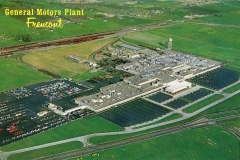 Aerial view of the General Motors Plant in Fremont. The facility is large and surrounded by green fields, with numerous cars parked in adjacent lots. Roads and a railway line run alongside the plant.