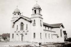 A historic black and white photo of Holy Ghost Catholic Church in Centerville, California, showcasing its ornate architecture with multiple domes, detailed facades, and a cross on top. A water tower is visible in the background.