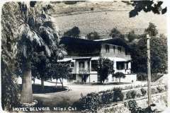 A black and white vintage photo of Hotel Belvoir in Niles, California. The building is surrounded by trees with hills in the background. Palm trees and a well-maintained garden are visible in the foreground. A sign reads Hotel Belvoir Niles Cal.