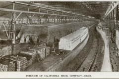 Interior of the California Brick Company in Niles, showing workers standing beside large kilns and stacks of bricks. The warehouse is spacious with high ceilings and railway tracks running alongside the kiln.