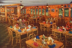 International Kitchen, Niles, California. A rustic dining room with wooden tables and chairs, set with colorful napkins and floral centerpieces. The room is warmly lit, and the walls are adorned with plates and large windows overlooking greenery.