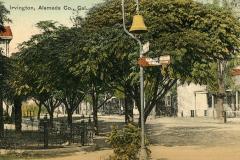 A vintage postcard depicts a street scene in Irvington, Alameda County, California. Large trees line the road, and a street sign points toward Mission San Jose. A distinctive bell is mounted on a pole in the foreground.