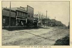 Black-and-white vintage photo of Main Street in Niles, California, facing west. The street is lined with early 20th-century storefronts and bare trees. A few signs and telephone poles are visible, along with a dirt road extending into the distance.