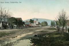 A vintage postcard depicts Main Street in Centerville, California. The dirt road is lined with horse-drawn carriages and rustic buildings. A church with a steeple stands in the background, and a mountain looms in the distance.