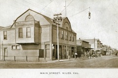 Sepia-toned vintage photo of Main Street, Niles, Cal. showing early 20th-century buildings and a dirt road. A horse-drawn carriage is visible near storefronts, and electric cables hang above the street.
