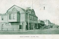 Vintage photograph of Main Street, Niles, California. It features historic buildings with a sign for a lodge, horse-drawn carriages, and people on the sidewalk. The scene captures early 20th-century architecture and a streetlight hanging overhead.