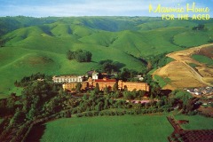 Aerial view of Masonic Home for the Aged, surrounded by lush green hills. A large building complex is nestled among trees, with open fields and a winding road nearby. Bright yellow text in the sky reads Masonic Home for the Aged.