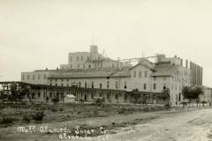 Black and white photo of a large industrial factory building marked Alvarado Sugar Co. and Alameda Sugar. It has multiple stories, smokestacks, and visible piping. Surrounding the building is an open landscape with sparse vegetation.