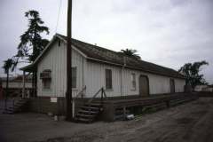 Niles, California, May 1979. A weathered, wooden warehouse with a sloped roof and two small staircases on either side, set against a cloudy sky. A large tree and utility pole are visible nearby. The building has a ramp along one side and is surrounded by dirt and pavement.