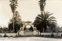 Vintage black and white photo of St. Marys School for Girls entrance with a decorative archway, tall palm trees on either side, and a cross atop the arch. A paved driveway leads through the open gate. The words Main Entrance are handwritten at the bottom.