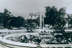 Stanford Ranch Fountain, Warm Springs, Alameda Co. California. A vintage photograph of a circular garden pond with water lilies and a fountain. Trees and plants surround the pond, with a grand building visible in the background. The setting is serene and well-maintained, suggesting a historical estate or park.