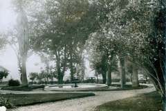 Stanford Ranch Gardens, Warm Springs, Alameda Co. A black and white photo of a garden featuring a circular fountain surrounded by trees and a gravel path. In the background, there is a vintage car parked on the left and a small building on the right.