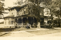 A vintage photo of a two-story Victorian-style house with a wraparound porch, surrounded by trees. A person is sitting on the front steps, and the house is situated on a grassy lawn.