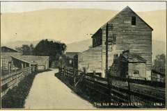 Black and white photo of The Old Mill in Niles, California. The mill is a large wooden structure beside a dirt road lined with wooden fences. Trees and hills are visible in the background.