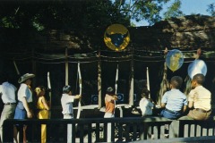Children and adults gather at the Frontier Village archery range in San Jose, California. Some kids aim bows at targets while others watch. Balloons and a sign with a bulls head are visible, surrounded by trees and rocky backdrops.