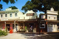 A colorful vintage street scene in California shows people walking past old-fashioned storefronts, including a sweet shop, a paddle shop, a glass shop, and a bank, much like the charm once found at San Jose’s famous Frontier Village.
