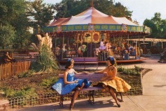 Two women in colorful dresses and bows sit on a bench, holding hands, with San Jose’s vibrant Frontier Village carousel and riders in the background. Green trees and a clear sky complete the cheerful California scene.
