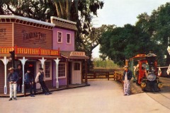 A colorful old-west style trading post and candy store stand on the left at Frontier Village in San Jose, California, with people dressed in cowboy attire. On the right, a small bright train marked Rail Road approaches along tracks, amid trees and wooden fencing.