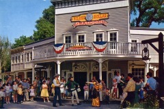 A crowd gathers outside the Silver Dollar Saloon at Frontier Village in San Jose, California—a Western-style building adorned with red, white, and blue bunting. Performers in period costumes entertain families and children on a sunny day.