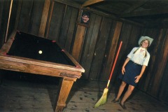 A woman in a cowboy hat stands barefoot beside a broom and pool table with a single ball, inside a wooden-paneled room with slanted floors. A framed photo hangs on the back wall at Frontier Village in San Jose, California.