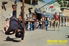 A cowboy performer falls during a stunt show in front of an Old West-style building at Frontier Village amusement park in San Jose, California. Onlookers, including children, watch the action as a large Mexican flag waves in the background.