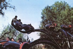 People ride a vintage amusement park ride with black and red cars, curving among green trees at Frontier Village in San Jose, California. Under the clear blue sky, some riders appear excited or smiling.