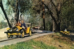 Two people smile as they ride in a yellow vintage-style car on a guided track through a wooded area at San Joses Frontier Village, while another yellow car follows behind. The scene is bright and outdoors.