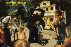 Children and adults gather around a person in a bear costume at Frontier Village amusement park in San Jose, California. The bear wears a hat, with a colorful building and a cowboy visible in the background.