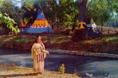 A woman in Native American-style dress stands by a stream at Frontier Village in San Jose, California, holding a decorated staff. Behind her are three colorful tipis and trees, with ducks swimming in the water at the front of the image.