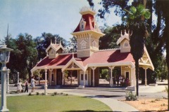 A vintage photo of a Victorian-style park pavilion with a red roof, ornate trim, and central clock tower, surrounded by trees at Frontier Village in San Jose. Several people are gathered near the building and on the walkway.