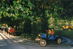 People ride colorful vintage-style cars along a shaded track surrounded by lush green foliage at Frontier Village in San Jose, California. Two children sit in a blue car in the foreground, while others wait in line or drive cars in the background.