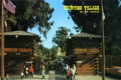 Entrance to Frontier Village in San Jose, California, with two large wooden gates, people walking, an American flag, tall trees, and a vintage orange car to the right. A bright sign reads “FRONTIER VILLAGE.”.