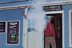 A man dressed as a cowboy stands in the doorway of the Marshal’s Office at Frontier Village in San Jose, California, aiming a smoking revolver toward the camera. Signs and a blue wooden building are visible.