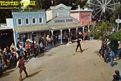 A crowd watches a staged gunfight between two cowboys in front of Old West-style buildings at Frontier Village amusement park in San Jose, California, with a ferris wheel visible in the background.
