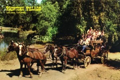 A vintage stagecoach pulled by four horses carries a group of people through a wooded area at Frontier Village in San Jose, California, under bright sunlight.