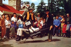 A crowd watches as two men in cowboy costumes wheel a person tied to a stretcher labeled Lightning Express outside a Western-style building at Frontier Village in San Jose, California; children and adults appear entertained and curious.
