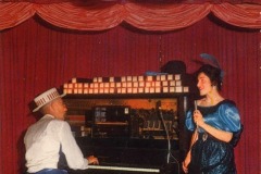 A man in a striped hat plays piano on stage at Frontier Village in San Jose, California, under a Silver Dollar sign, while a woman in a blue dress and feathered hat sings beside him. Red curtains hang behind them.