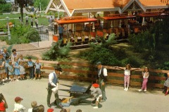 A crowd watches as three performers in cowboy attire act out a scene near a vintage open-air train at Frontier Village in San Jose, California, with children and adults gathered around a wooden fence in a park setting.